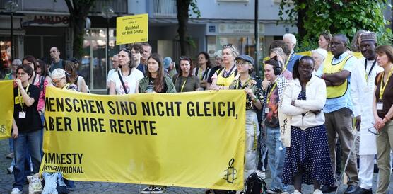 Das Foto zeigt eine Gruppe von circa 30 Personen, die auf einem Platz stehen und einer Rede zuhören. Einige halten ein Banner vor sich, auf dem steht: "Menschen sind nicht gleich, aber ihre Rechte".