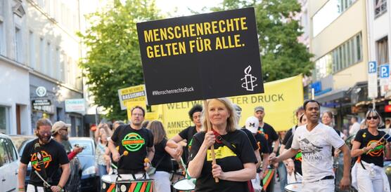 Das Foto zeigt eine Frau an der Spitze eines Demonstrationszuges. Sie hält ein Schild hoch mit Amnesty-Logo und dem Slogan "Menschenrechte gelten für alle!". Hinter ihr spielt eine Trommelgruppe.