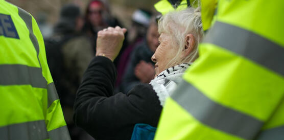 Das Foto zeigt eine ältere Frau mit Palästinensertuch inmitten von uniformierten Sicherheitskräften. Sie reckt die linke Faust hoch.