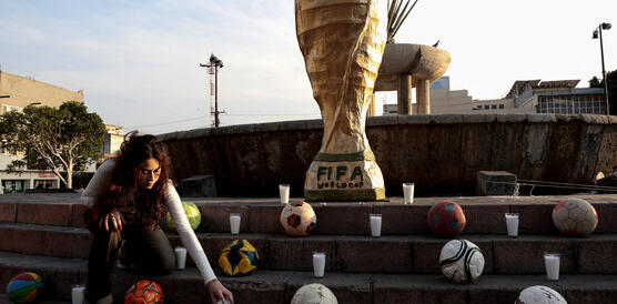 Kunstaktion in Mexiko: Eine Studentin stellt eine Kerze zu einer großen FIFA-WM-Pokal-Nachbildung, die an vermisste Personen erinnert und von Fußbällen umgeben ist.