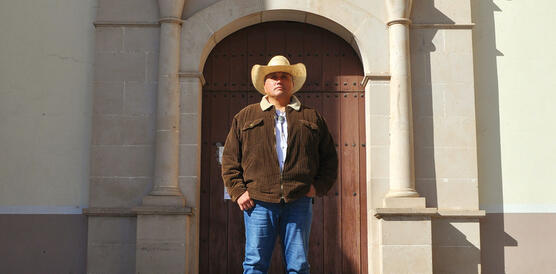Ein indigener Mann trägt Jeans, Jacke und einen Cowboyhut, er steht vor der Front eines alten Gebäudes.
