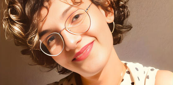 Eine junge Frau mit Locken in einer Bluse hält einen Kaffeebecher in der rechten Hand und lächelt.
