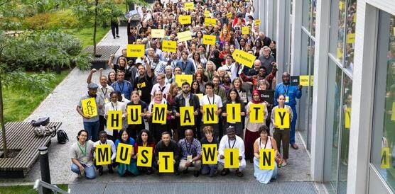 Gruppenfoto mit zig Menschen neben einem Gebäude. Einige halten Schilder hoch. Die Personen vorne halten Tafeln mit Buchstaben hoch, die die Slogan bilden: "Humanity must win."