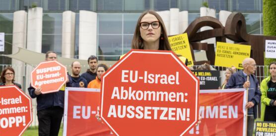 Menschen bei Protestaktion vor dem Kanzleramt in Berlin mit Transparenten und Schildern. Eine Frau steht im Vordergrund. Auf ihrem Schild steht: "EU-Israel-Abkommen aussetzen!"