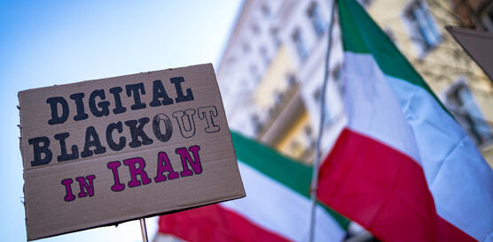 Eine Nahaufnahme eines Pappschildes mit der handgeschriebenen Aufschrift „DIGITAL BLACKOUT IN IRAN“, gehalten vor einem unscharfen Hintergrund mit iranischen Flaggen und der Fassade eines europäischen Gebäudes während einer Demonstration.