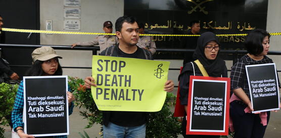Vier Demonstrierende stehen nebeneinander vor dem Gebäude der saudi-arabischen Botschaft in Jakarta und halten Protestschilder in die Kamera. Eine Person in der Mitte hält ein großes, leuchtend gelbes Schild mit der Aufschrift „STOP DEATH PENALTY“ und dem Logo von Amnesty International.