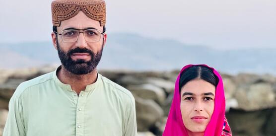 Das Foto zeigt Dad Shah und Fozia Baloc nebeneinander in einer Landschaft stehen und leicht in die Kamera lächelnd. Sie tragen traditionelle Kleidung.