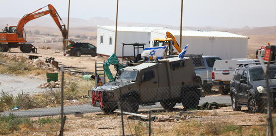Ein gepanzertes Militärfahrzeug mit zwei israelischen Flaggen steht neben Baggern, weißen Containergebäuden und Fahrzeugen in einer kargen Wüstenlandschaft.