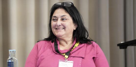 Das Foto zeigt Shenilla Mohamed lächelnd auf einem Podium sitzend. Sie schaut leicht zur linken Bildseite. Ihre Brille hat sie in ihre Haare gesteckt.