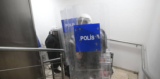 Mehrere Bereitschaftspolizisten mit weißen Helmen, Visieren und dunkler Schutzkleidung halten große durchsichtige Schutzschilde mit der Aufschrift „POLİS" in einem Treppenhaus, auf dessen Boden grüne Glasscherben liegen.