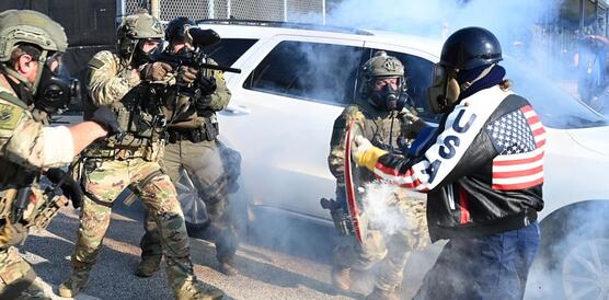 Bewaffnete Polizeikräfte gehen auf eine Person mit einer Gasmaske, einem Schild und einer Motorradjacke auf der "USA" steht zu.