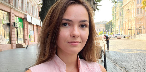 Eine junge ukrainische Frau mit langen Haaren steht auf einer Straße vor einem Baum und trägt eine leichte Bluse.
