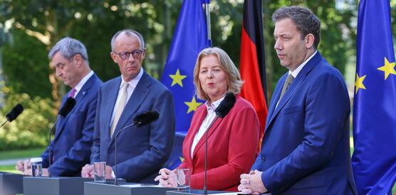 Markus Söder, Friedrich Merz, Bärbel Bas und Lars Klingbeil stehen nebeneinander im Freien vor kleine Podien mit Mikros. Hinter ihnen steht eine Deutschland-Flagge und zwei Flaggen der Europäischen Union.