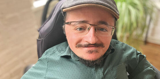 Ein Mann mit brillB und Schnurrbart trägt Hemd und Schiebermütze, er sitzt in einem Rollstuhl in einem Büro, im Hintergrund Topfpflanzen.