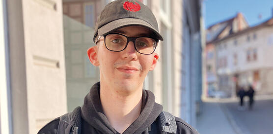 Eine junge Person trägt Baseball-Mütze und Brille und steht auf einer Straße.