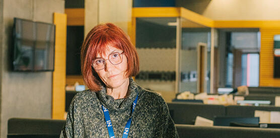 Eine Redakteurin, Frau in mittlerem Alter mit kinnlagem Haar, steht im Redaktionsraum eines TV-Senders, sie trägt eine Bluse und hält ein Blatt Papier in der Hand. 