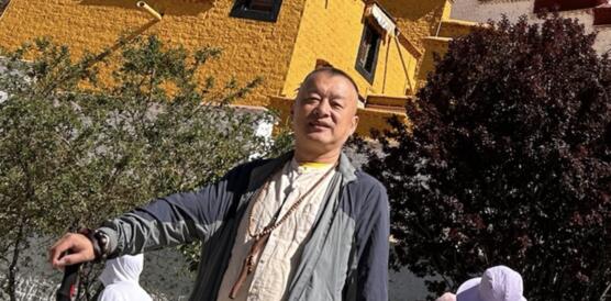 Das Foto zeigt Zhang Yi vor einem Gebäude stehen. Er blickt in die Kamera und hat seine rechte Hand auf einer Art Wanderstock gestützt.