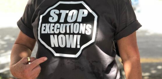 Aufnahme einer Person an einem sonnigen Tag, die mit ihrer rechten Hand auf ihr T-Shirt zeigt, auf dem steht: "Stob executions now!"