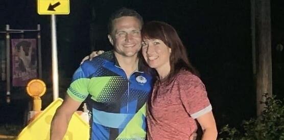 Das Foto zeigt Andriy Shepitsen, der in Sportkleidung hinter einem Fahrrad steht und in die Kamera lächelt. Neben ihm steht eine Frau, die ebenfalls lächelt. Sie umarmen sich gegenseitig. Das Foto wurde abends oder nachts aufgenommen vor einem Verkehrsschild.