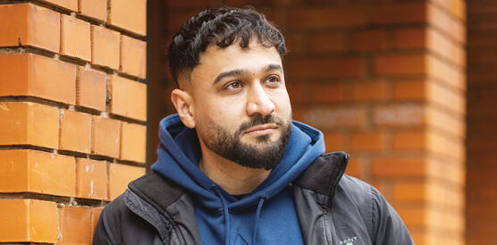 Junger Mann in adidas-Hoodie und Kurzjacke lehnt vor einem gebäude aus Backsteinziegeln.