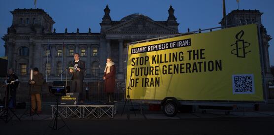 Das Bild zeigt den Deutschen Bundestag bei Nacht, davor steht ein Schild auf einem Wagen mit dem Schriftzug "Stop killing the future generation of Iran". Daneben stehen mehrere Personen, von denen eine in ein Mikro spricht.