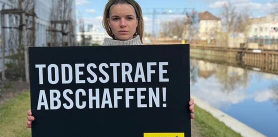 Das Foto zeigt eine junge Frau. Sie hält ein Schild mit Amnesty-Logo vor sich. Auf dem Schild steht: "Todesstrafe abschaffen". Links neben ihr steht ein Gebäude. Rechts von ihr ist ein Gewässer.