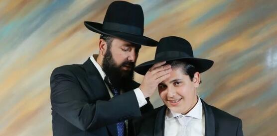 Rabbi Yitzak Biton (links) und sein Sohn stehen nebeneinander, mutmaßlich in einem Fotostudio. Sie tragen Anzüge und Hüte. Yitzak Biton legt seinem lächelnden Sohn die Hand seine linke Hand auf die Stirn.
