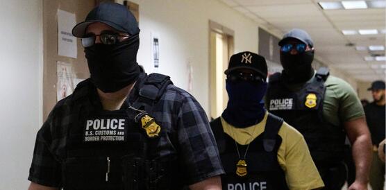 Das Foto zeigt drei Männer mit Basecap, Sonnernbrillen und Skimasken, die ihr halbes Gesicht bis unter die Augen bedecken. Sie gehen hintereinander in einem Flur. Sie tragen Schutzwesten über ihrer zivilen Kleidung und unter anderem eine Pistole im Holster.