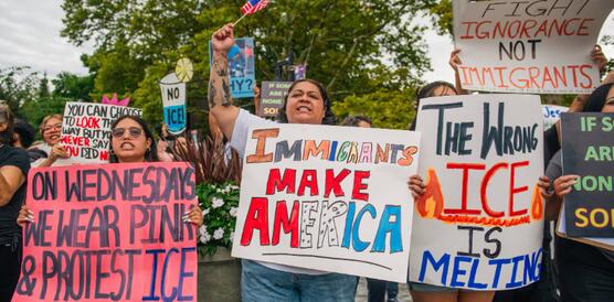 Das Foto zeigt mehrere Frauen bei einer Demonstration. Sie halten Schilder hoch. Auf einem steht: "The wrong ICE is melting". Auf einem anderen: "Immigrants make America".