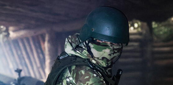 Das Foto zeigt einen maskierten Soldaten mit Helm und Schutzweste in einem Unterstand. Hinter ihm steht ein Geschützt.