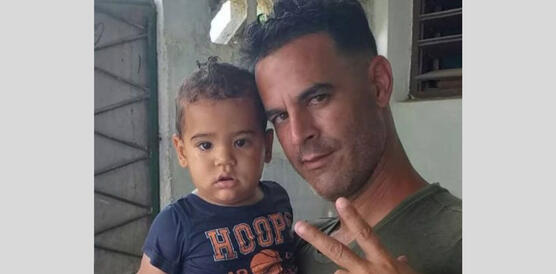 Roberto Pérez Fonseca hält ein kleines Kind in seinem rechten Arm. Beide blicken in die Kamera. Fonseca hält den den Zeige- und Mittelfinger seiner linken Hand gespreizt nach oben.