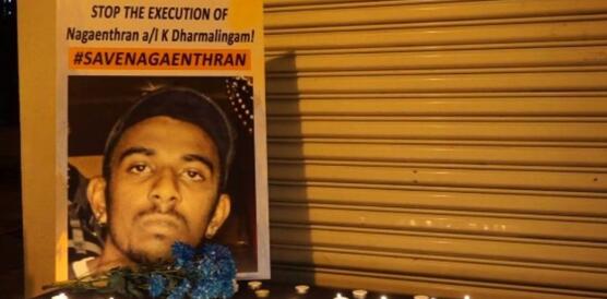 Ein Plakat mit einem Porträtfoto von Nagaenthran Dharmalingam steht auf einem Tisch vor einem Wellblechtor. Vor dem Plakat stehen Kerzen. Auf dem Plakat steht: "Stop the execution of Nagaenthran a/I K Dharmalingam!"