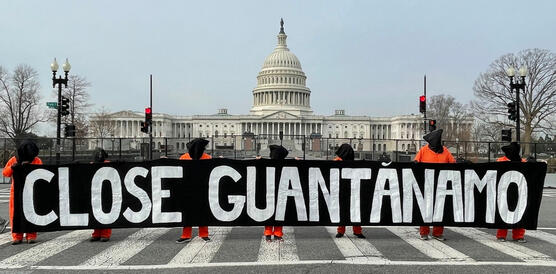 Mehrere Personen in Gefangenenkleidung und mit Säcken über den Kopf halten auf einer Straße ein großes Banner vor sich mit der Aufschrift: "Close Guantanamo". Im Hintergrund steht ein großes Geäude: Das Kapitol.