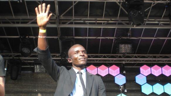 Emmanuel Odhiambo Nyambwa, in schwarzem Anzug, winkt mit ausgestrecktem Arm auf einer Bühne