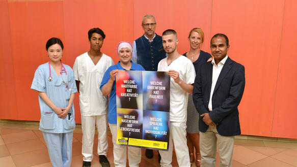 Mitarbeiterinnen und Mitarbeiter des Klinikums München halten ein Plakat der Kampagne "Nimm Rassismus persönlich" hoch.