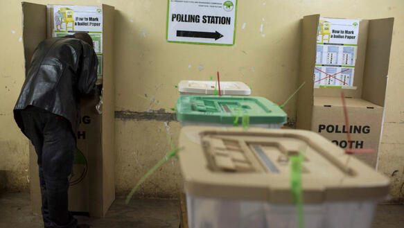 Ein Mann beugt sich in einer Wahlkabine nach vorne, um seine Stimme abzugeben, im Vordergrund stehen Plastikwahlurnen