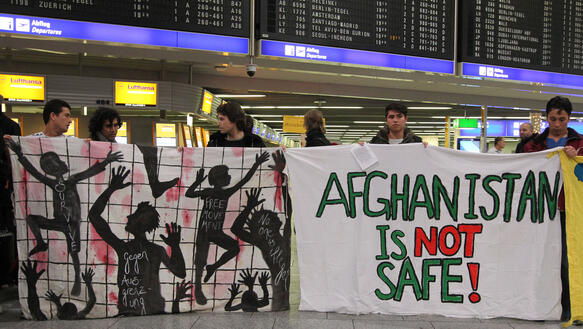 Mehrere junge Männer halten Transparente hoch mit Zeichnungen und dem Slogan "Afghanistan is not safe!", im Hintergrund ist eine Anzeigetafel zu sehen mit den Zeiten der Abflüge vom Flughafen
