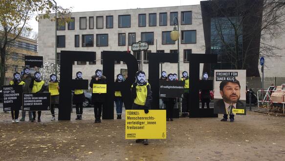 Mehrere Aktivistinnen und Aktivisten stehen neben vier großen Buchstaben, die das Wort "Free" bilden, und halten sich Papptafeln mit dem Porträtfoto von Taner Kılıç vor das Gesicht und halten Schilder mit Forderungen in den Händen 