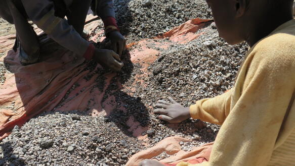 Zwei Kinder hocken auf dem Boden und wühlen mit den Händen durch Erde und Steine, die auf einer Plane ausgebreitet sind