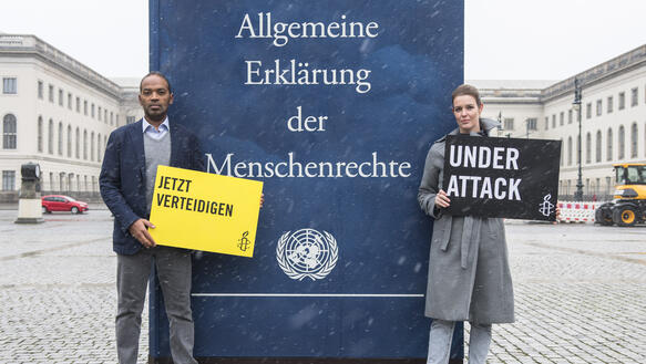 Auf einem verschneiten Platz steht ein schwarzer Mann im blauen Sakko und eine Frau im grauen Mantel um eine überdimensionale Ausgabe der Allgemeinen Erklärung der Menschenrechte. Er hält ein gelbes Schild mit der Aufschrift "Verteidigen“ und sie ein schwarzes Schild mit dem Spruch "Under Attack“.