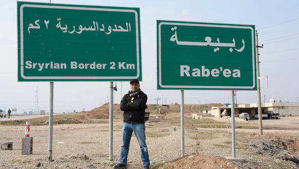 Mohammed Khamis steht vor zwei grünen Straßenschildern, auf denen "Syrian Border 2Km" und "Rabe'ea" auf Arabisch und Englisch steht