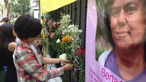 Menschen binden Blumen an einen Zaun, an dem auch ein Porträtfoto der Menschenrechtsverteidigerin Berta Cácares