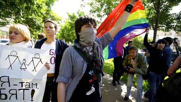 Eine junge vermummte Protestirende schwenkt im Zentrum des Bildes eine Regenbogenfahne in dei Luft, links von ihr halten zwei junge Frauen Protestplakate hoch und im Hintergrund sieht man weitere Protestierende und einen Fotojournalisten