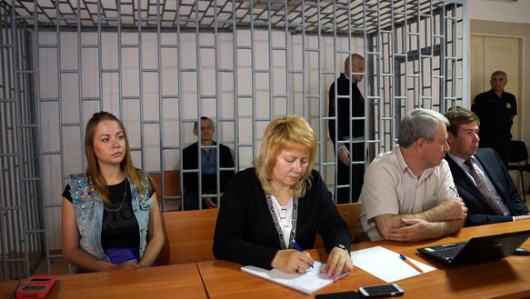 Klykh sitzt und Karpyuk steht in einem Käfig in einem Gerichtssaal, vor dem Käfig sitzen mehrere Personen an einem Tisch