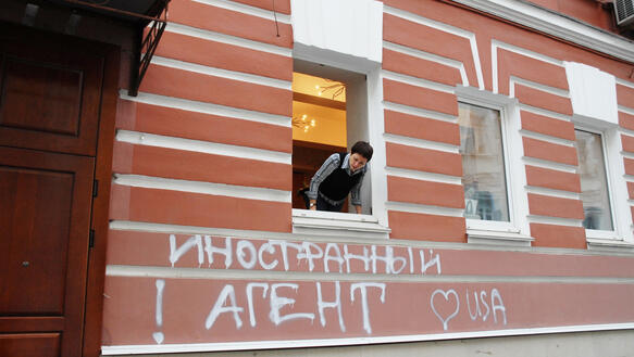 Ein Haus von außen, aus einem Fenster schaut eine Person, Graffiti auf russisch an der Wand: ausländischer Agent, ein Herz, USA