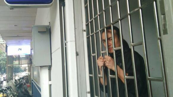 Zhen Jianghua ist hinter Gittern, seine Hände an den Gitterstäben. An der Außenwand ein Schild: Polizeiwache.