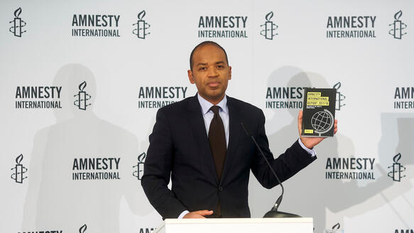 Ein Mann im Anzug steht hinter einem Rednerpult und hält ein schwarzes Buch mit dem Titel "Amnesty International Report 2017/18" hoch