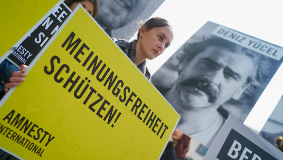 Eine junge Frau hält ein großes Schild vor sich mit der Aufschrift "Meinungsfreiheit schützen", im Hintergrund ist auf einem Plakat ein Porträtfoto von Deniz Yücel zu sehen
