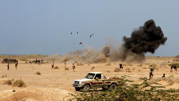 Nach einer Explosion steigt Rauch in einer Sandlandschaft auf, im Vordergrund sieht man bewaffnete Männer und einen Pick-Up