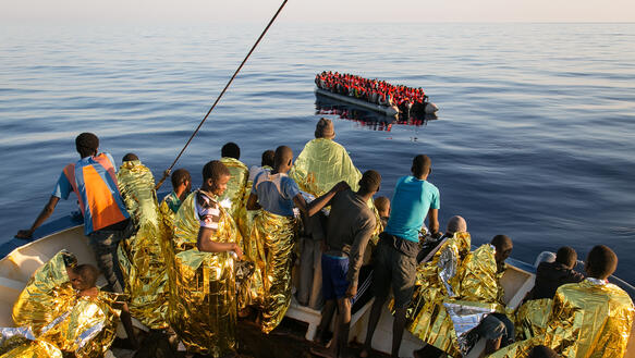 Eine Gruppe Personen in Rettungsdecken sammelt sich im Bug eines Schiffs, um zu beobachten wie ein Schlauchboot längsseits genommen wird.